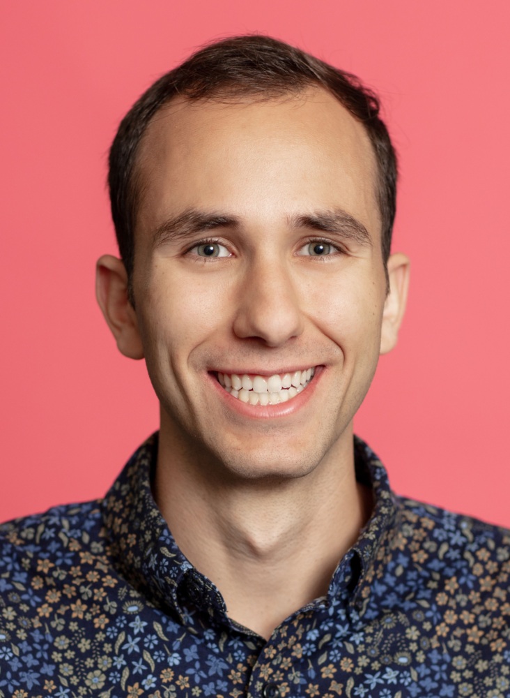 A man with short brown hair smiles at the camera. He wears a navy blue shirt with a floral pattern, and a pink background is behind him.